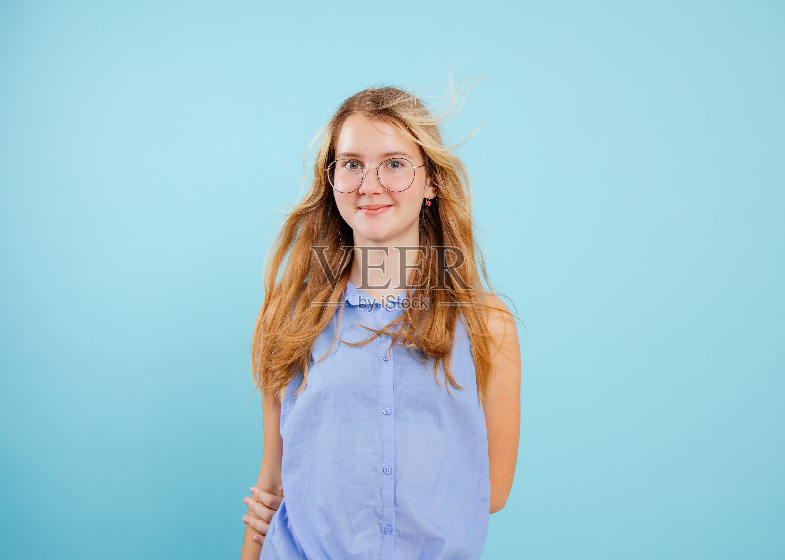 松散的头发十几岁的女孩站与手保持背后的空蓝色背景。肖像微笑学龄儿童在时尚的眼镜和无袖衬衫。青少年时尚，夏装照片摄影图片