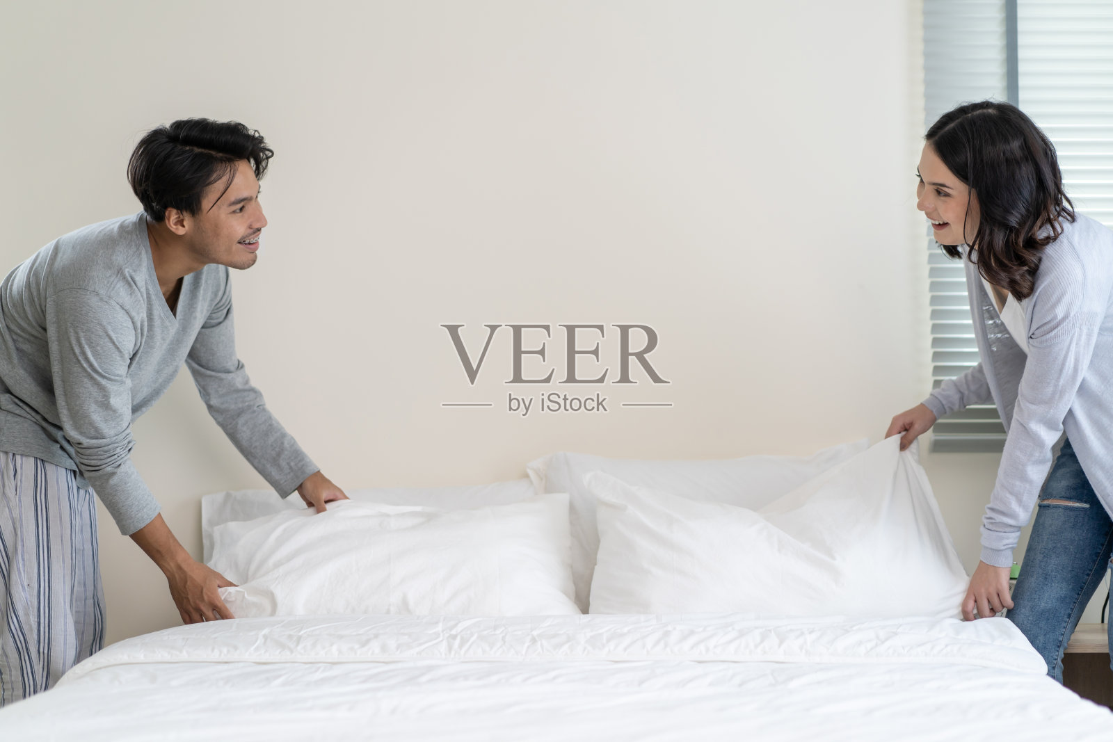 亚洲年轻夫妇早上一起起床后叠毛毯。有吸引力的新婚男女穿着睡衣在家里早早地从床上爬起来后感到幸福和放松。照片摄影图片