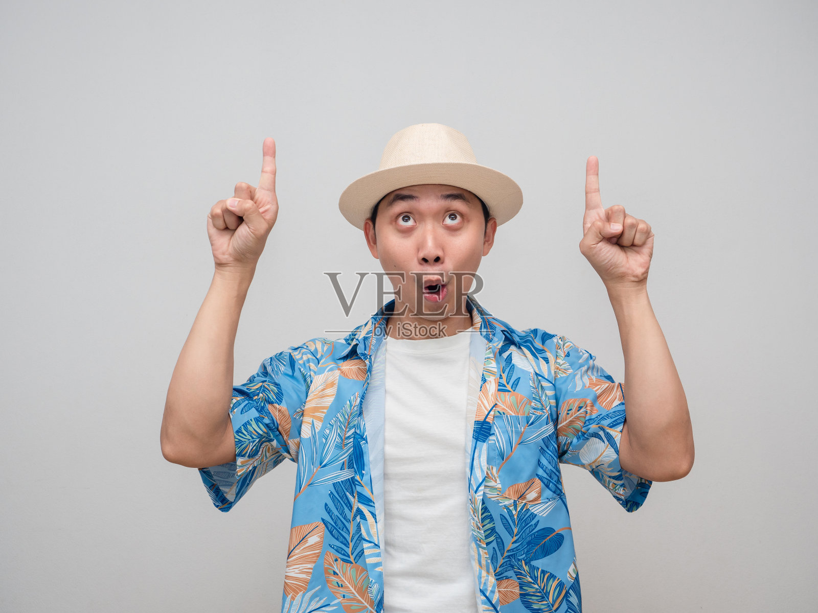 年轻人戴着帽子，穿着沙滩衬衫，感到兴奋，手势指着上面的手指照片摄影图片