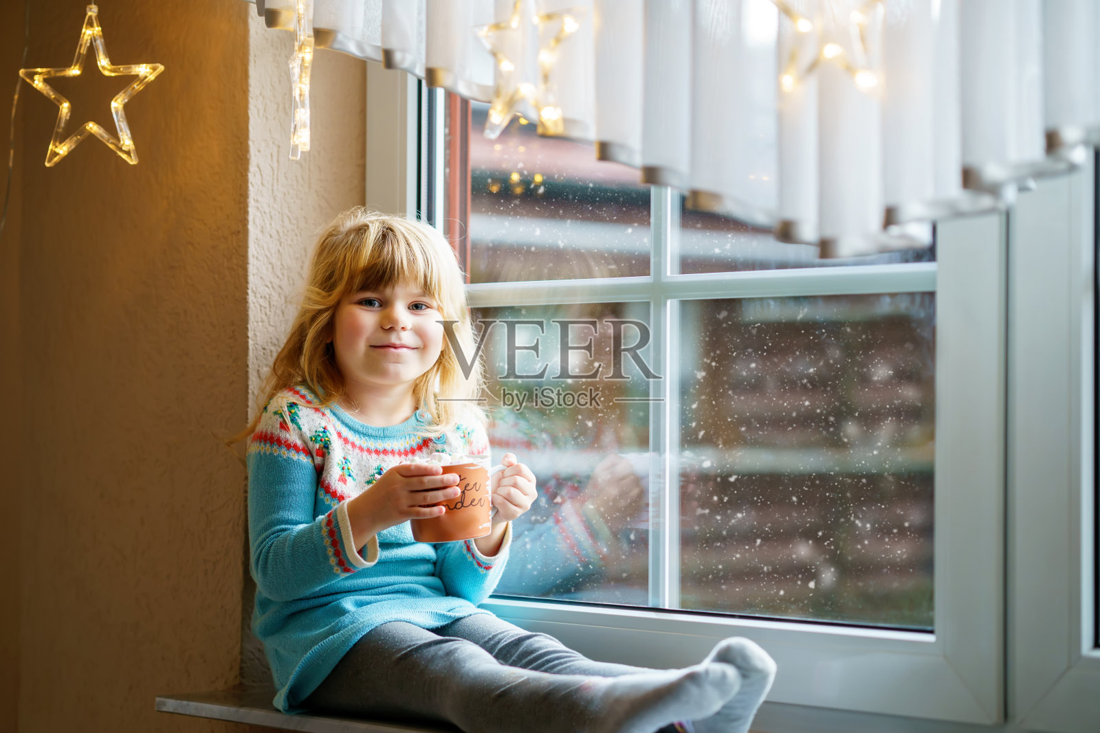 学龄前的小女孩拿着一杯热巧克力和棉花糖。快乐的孩子在冬天的圣诞灯下喝着甜可可。舒适的家庭庆祝圣诞节。照片摄影图片