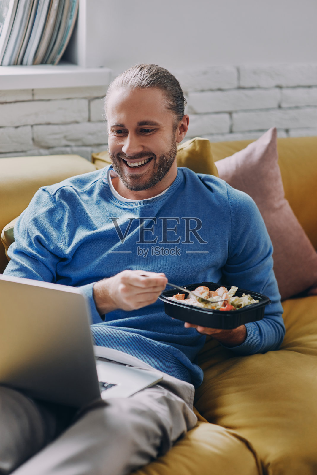 快乐的年轻人吃着午餐，坐在家里的沙发上看着笔记本电脑照片摄影图片