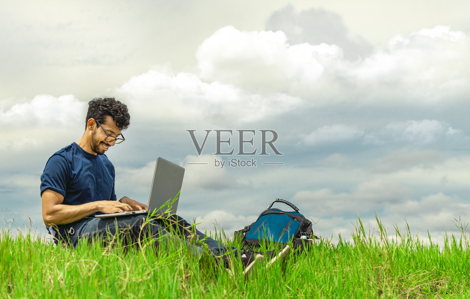 微笑的自由职业者坐在笔记本电脑和背包在领域。一个男人坐在草地上用他的笔记本电脑工作的肖像。自由职业者的概念，从现场工作照片摄影图片