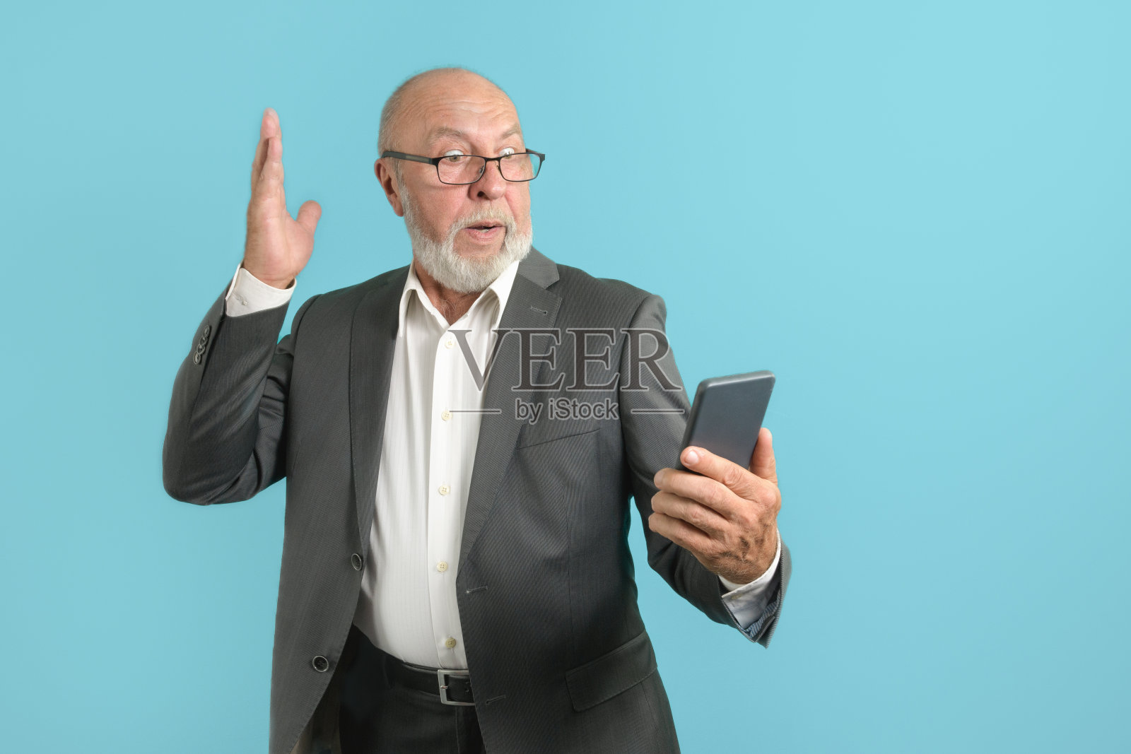 惊讶的消息。穿着经典西装的英俊老男人用手机隔离在蓝色背景的工作室肖像照片摄影图片
