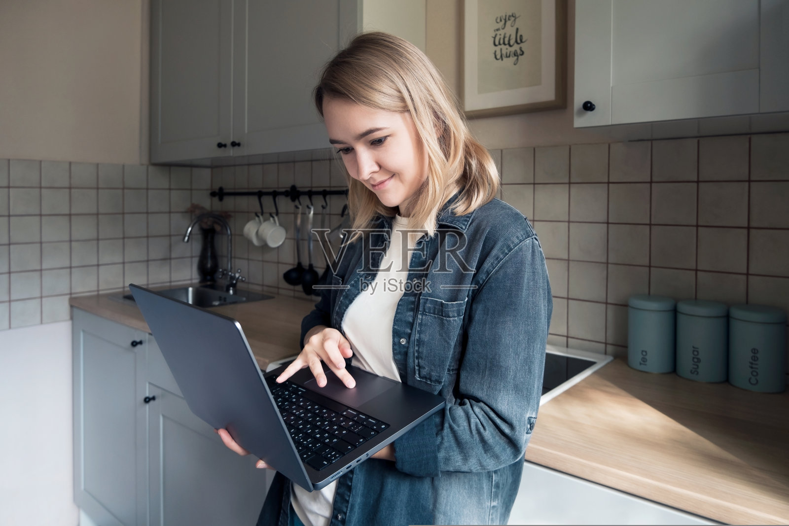 一个年轻的金发女孩拿着一台笔记本电脑站在厨房里，在家办公。远程工作的概念，家庭主妇的生活照片摄影图片