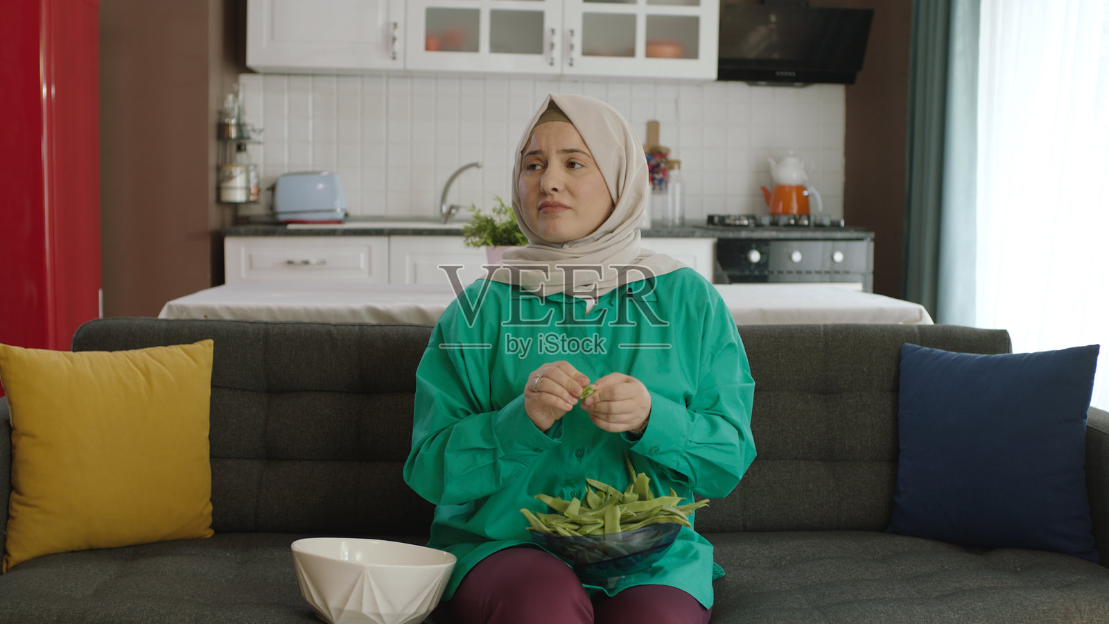 戴着头巾的年轻女子一边看电视一边切青豆。妇女准备食物的肖像。照片摄影图片