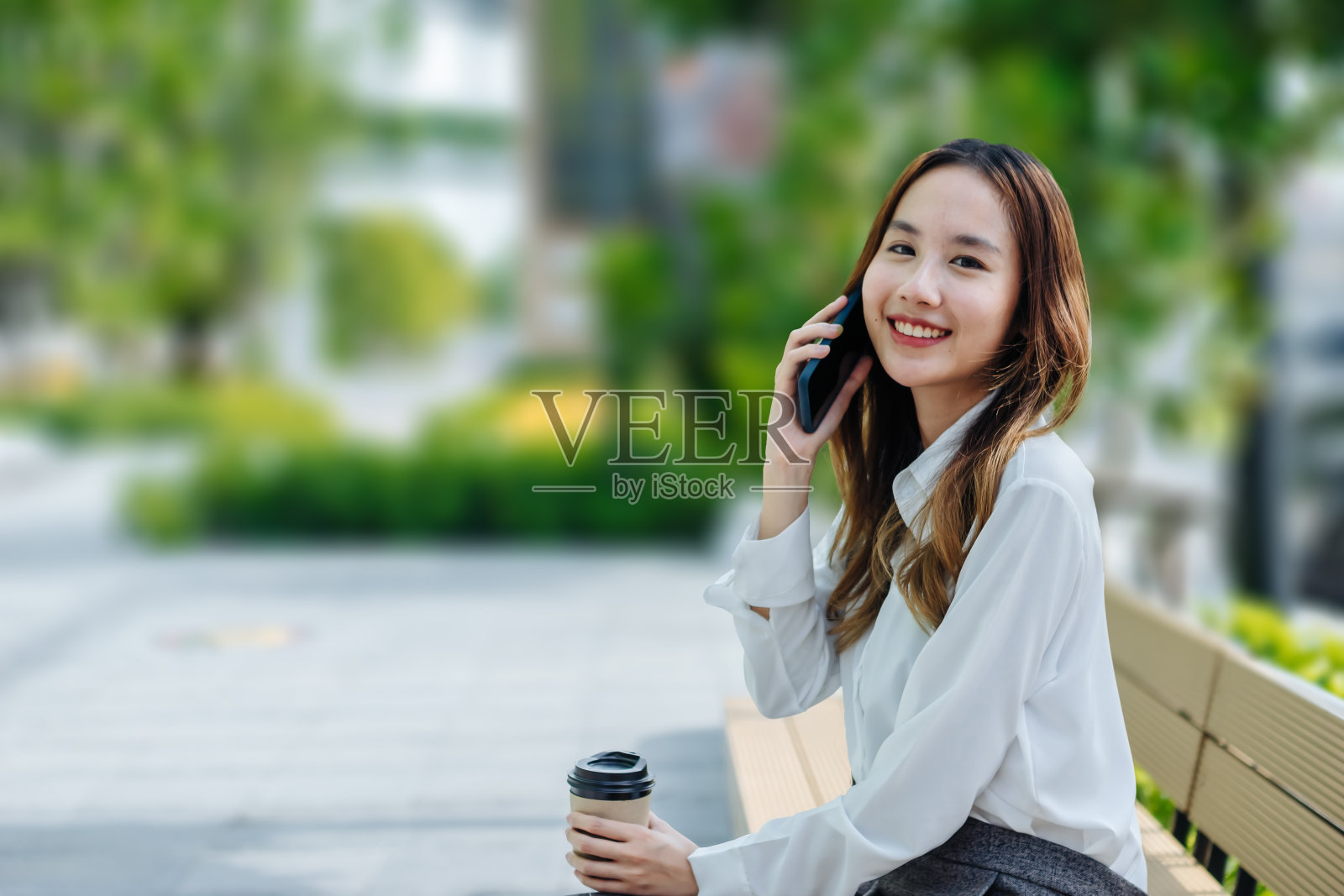 微笑着的亚洲卷发商务女性穿着时髦的衣服走在市中心的街道上，用着她的手机。漂亮的夏天女人在白衬衫走在街上看她的手机照片摄影图片