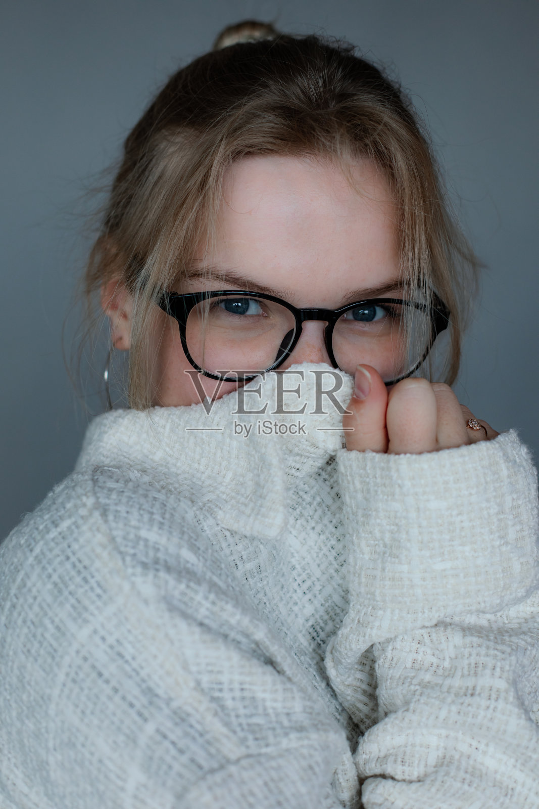 戴着黑色眼镜的年轻女子把鼻子和嘴藏在衣服下面，灰色背景，自由复制空间。舒适的白色毛衣，适合寒冷天气的衬衫。休闲时令服装，女性基本穿搭照片摄影图片