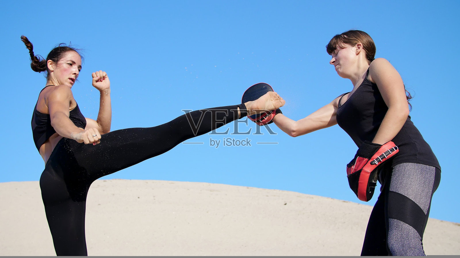 夏天，在烈日炎炎、蓝天下、荒芜的海滩上，两名身穿黑色健身服的年轻健美女子正穿着一双运动鞋，训练打架。慢动作照片摄影图片