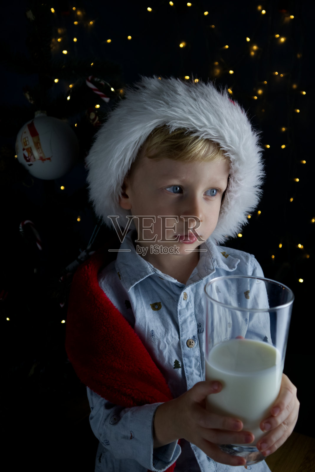 在圣诞树和彩灯的背景下，一个戴着圣诞老人帽的可爱男孩手里拿着一杯牛奶照片摄影图片