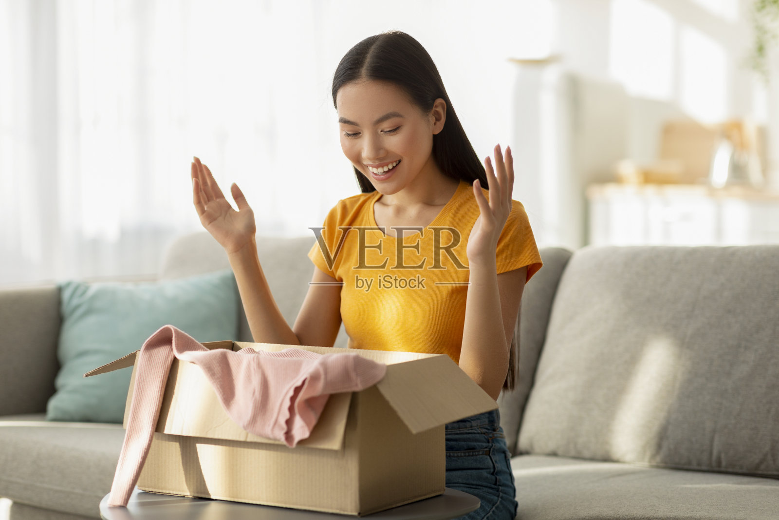 拆包的衣服。画像快乐的亚洲女士打开纸板箱，拿出新衬衫，坐在家里的沙发上照片摄影图片