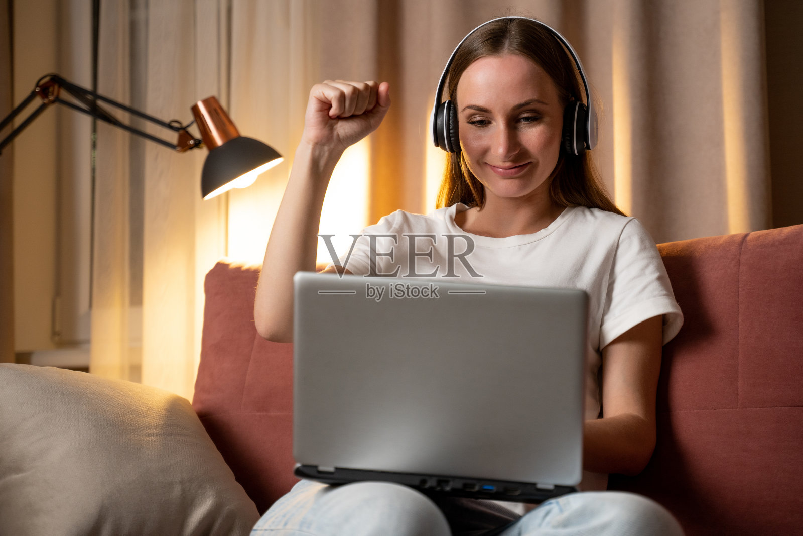 在现代化的公寓里，戴着耳机，举起双手，坐在舒适的沙发上使用笔记本电脑，满足的年轻学生为成功而高兴照片摄影图片