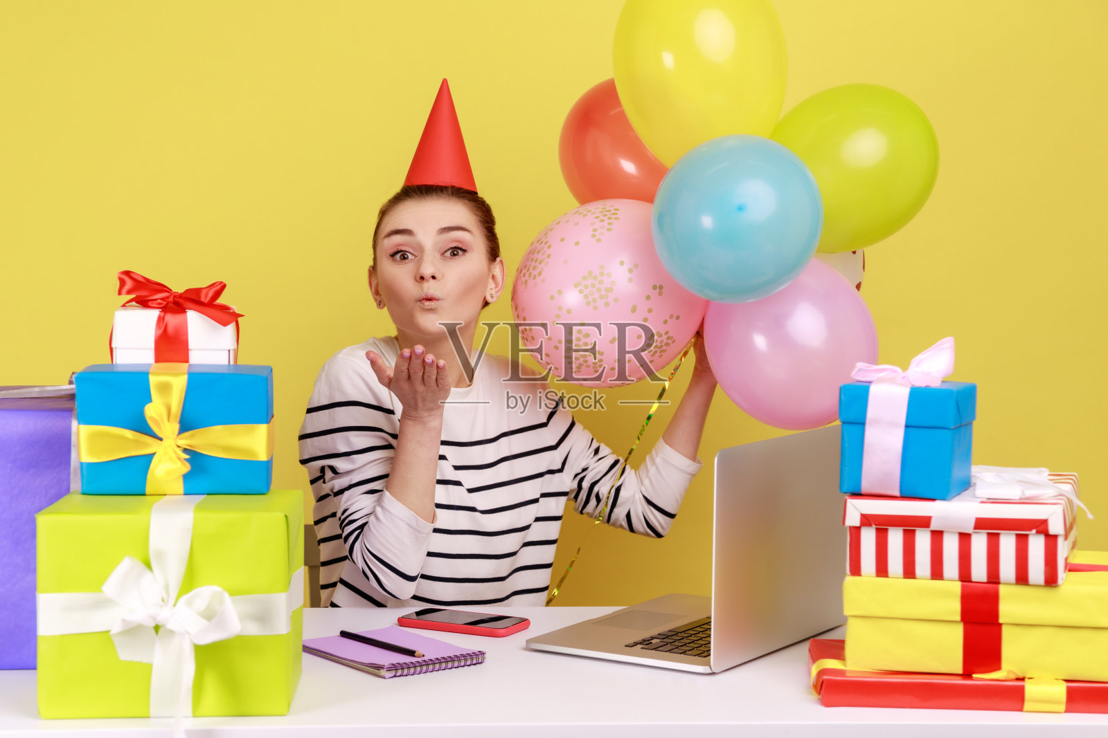 带着派对锥的女人坐在工作场所，周围是礼盒和气球，送上飞吻。照片摄影图片