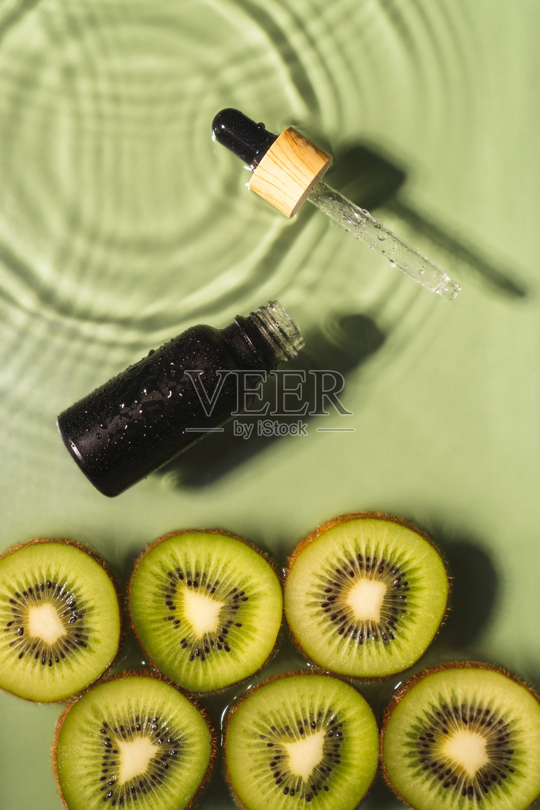 面部移液管精华黑色瓶，乳清美容产品附近的水果猕猴桃切片在水中飞溅新鲜透明。垂直照片摄影图片
