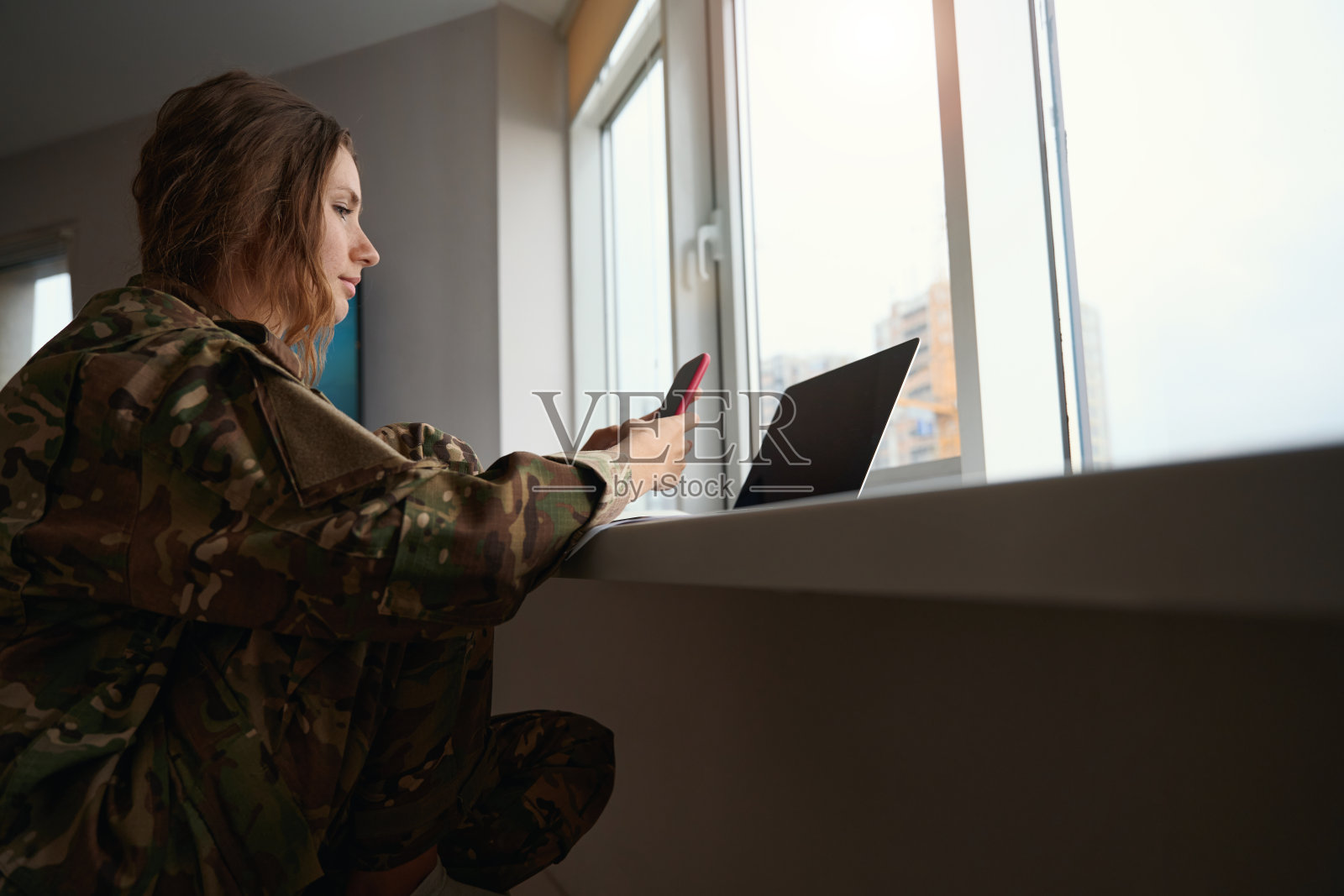 一名女军人坐在打开的笔记本电脑前，手里拿着手机照片摄影图片