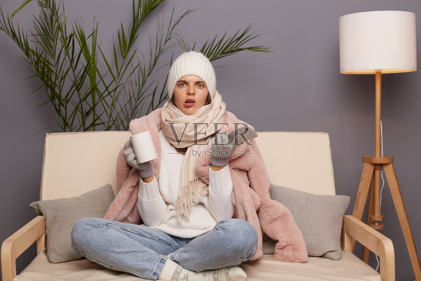 画像困惑年轻的成年妇女穿着温暖的衣服，裹着围巾坐在咳嗽杯热茶，困惑，使用智能手机。照片摄影图片