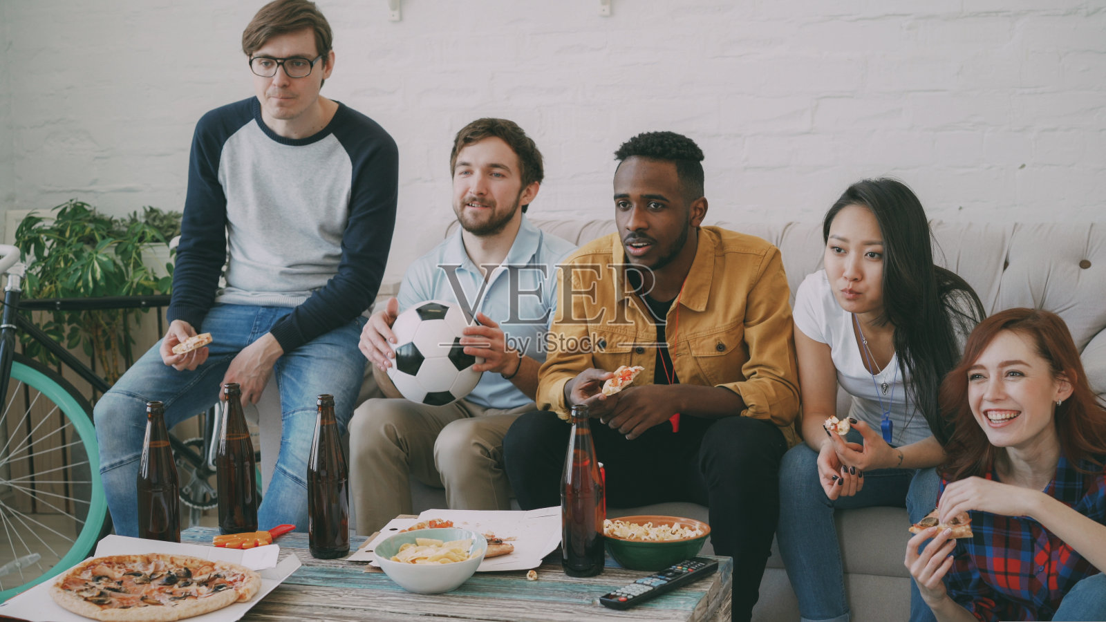 多民族的朋友，体育迷一起看电视上的足球冠军，在家里吃披萨，喝啤酒照片摄影图片
