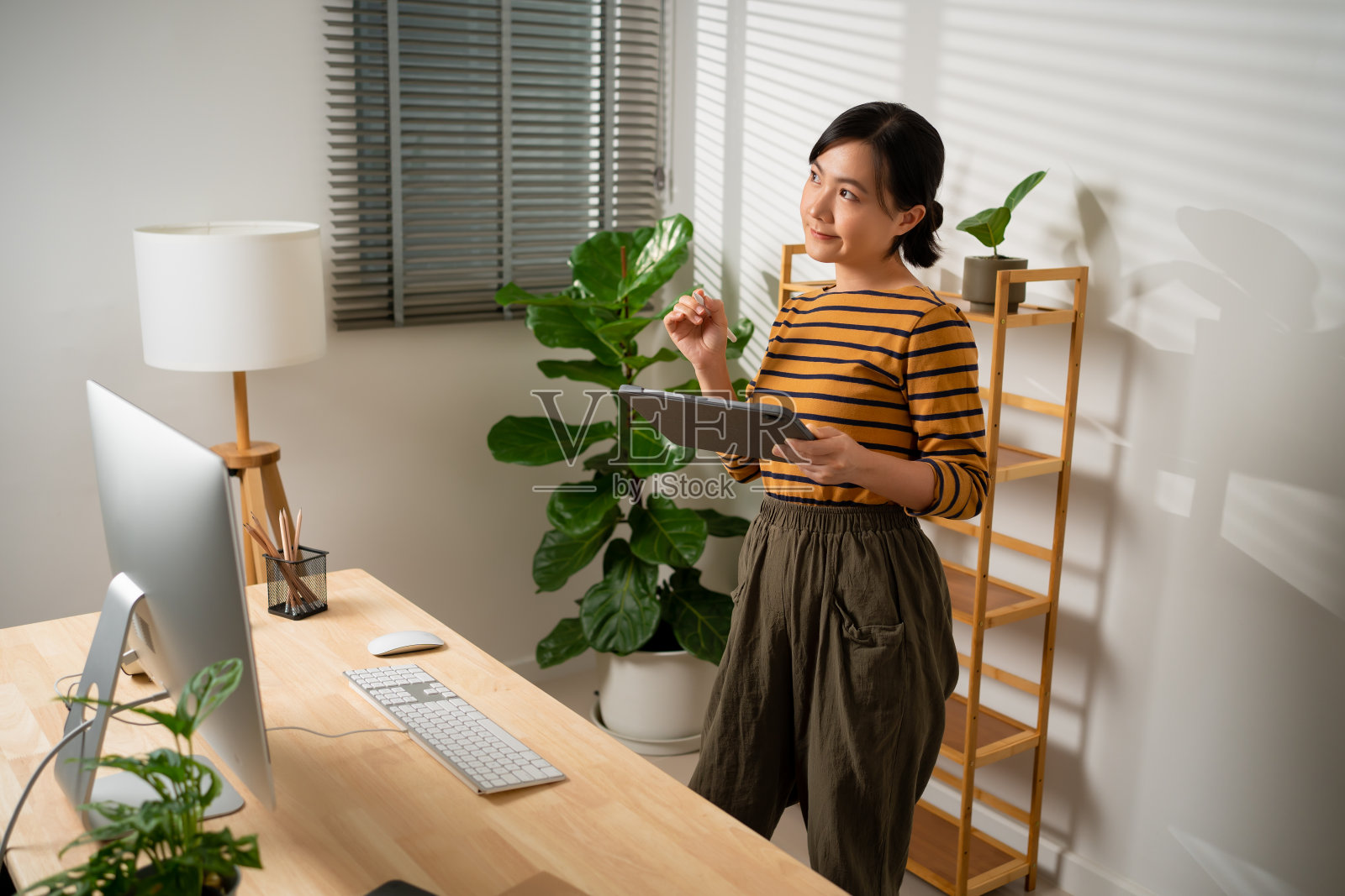 亚洲妇女手里拿着智能笔和平板电脑，站在家庭办公室的电脑前。照片摄影图片