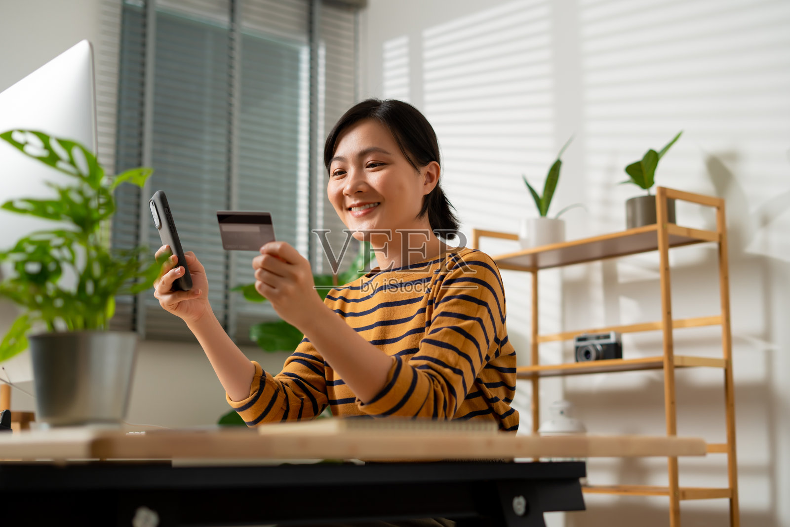 亚洲妇女拿着信用卡，用智能手机在家办公室网上购物。照片摄影图片
