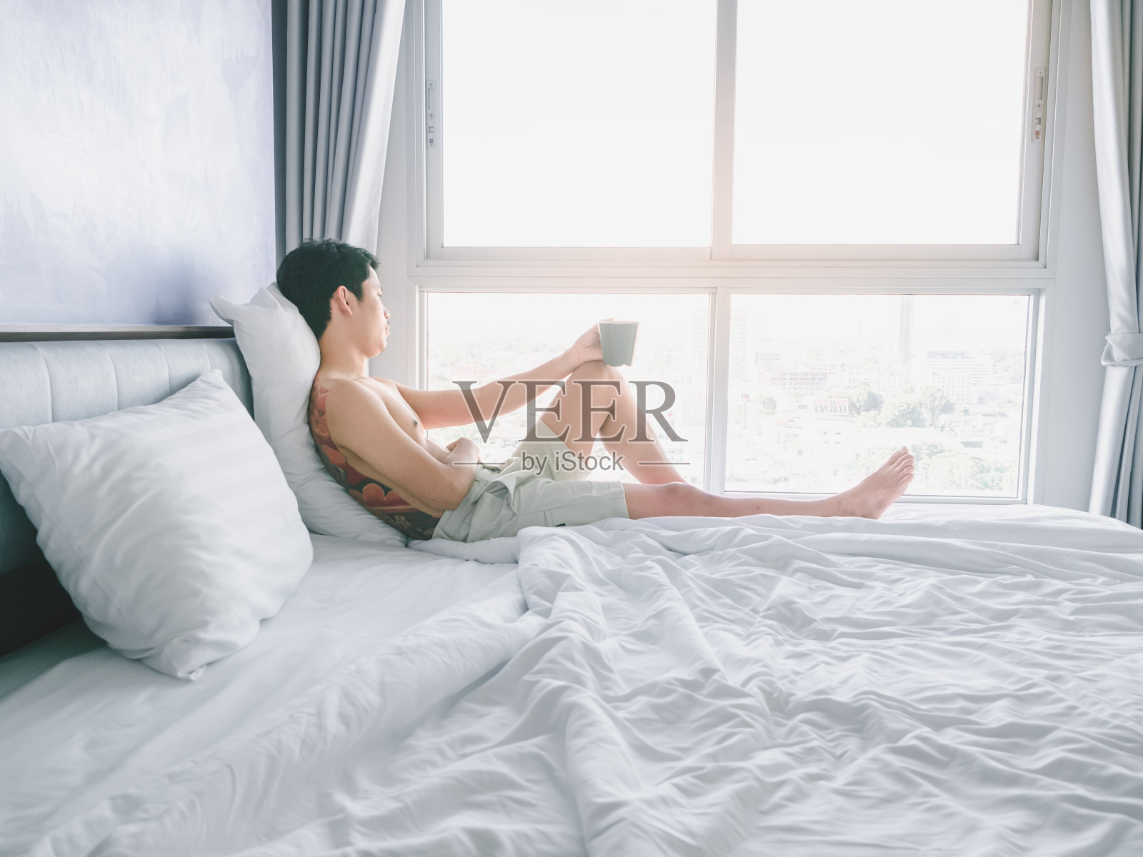 有纹身的亚洲青年，手拿咖啡杯放在床上，在晨曦中望向窗外照片摄影图片