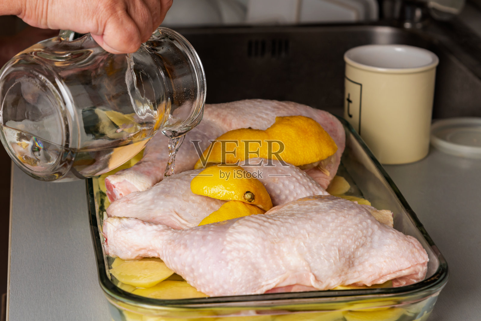 在家里的厨房里，烤盘里放着三只鸡腿，上面放着土豆、洋葱和柠檬角，一只手拿着水壶往鸡腿上浇水。照片摄影图片