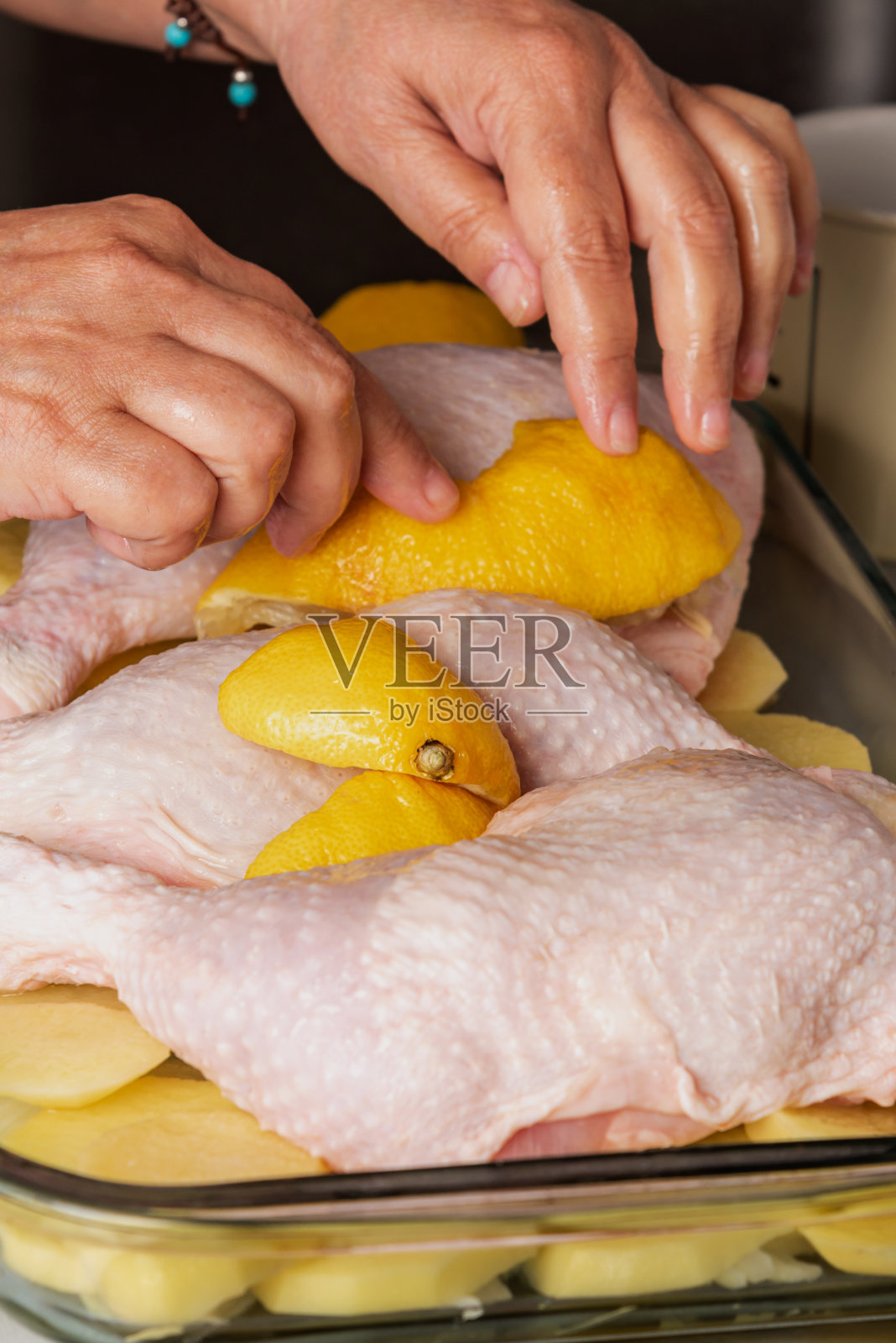 在家里的厨房里，烤盘里有三只鸡腿，上面放着土豆和洋葱碎，用手在鸡腿之间放一片柠檬。照片摄影图片