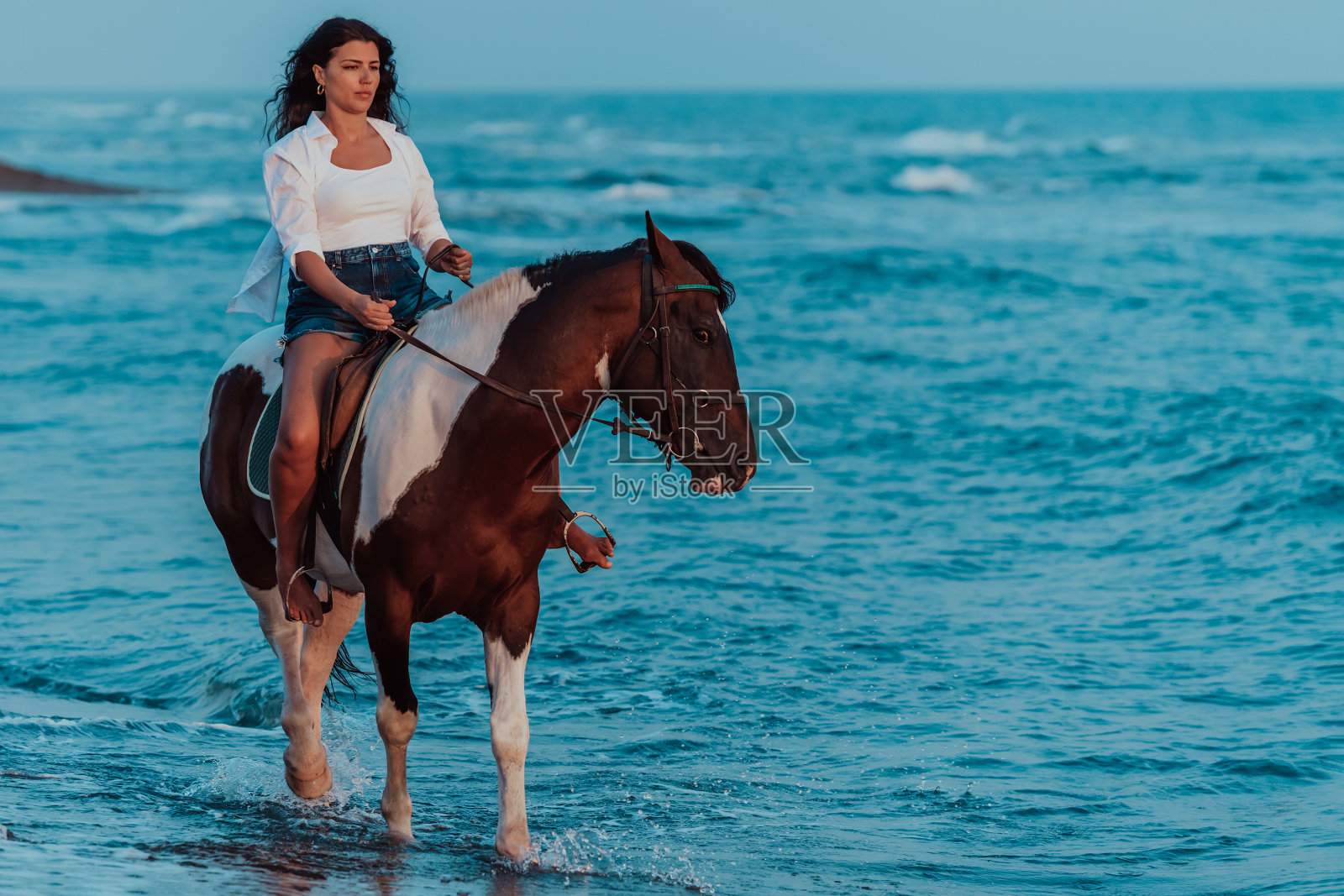 穿着夏装的女人喜欢在日落时分在美丽的沙滩上骑马。有选择性的重点照片摄影图片