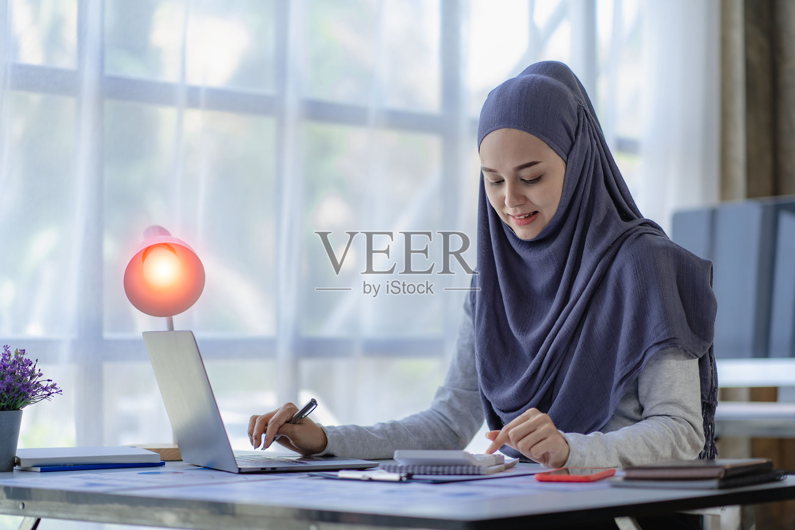 一位美丽的亚洲穆斯林妇女戴着头巾，在家里的客厅里用笔记本电脑做财务会计工作。照片摄影图片