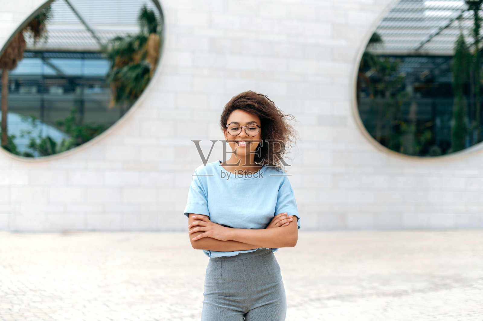 肖像正面优雅漂亮的非裔美国年轻女性卷发，戴眼镜，穿蓝色衬衫，站在户外双臂交叉，看着相机，微笑照片摄影图片