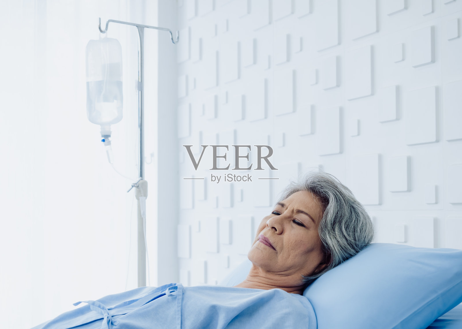 在现代化的白色病房里，白头发的亚洲老年女性病人睡在床上。一位老妇人生病了，躺在病床上，靠生理盐水滴液恢复。照片摄影图片