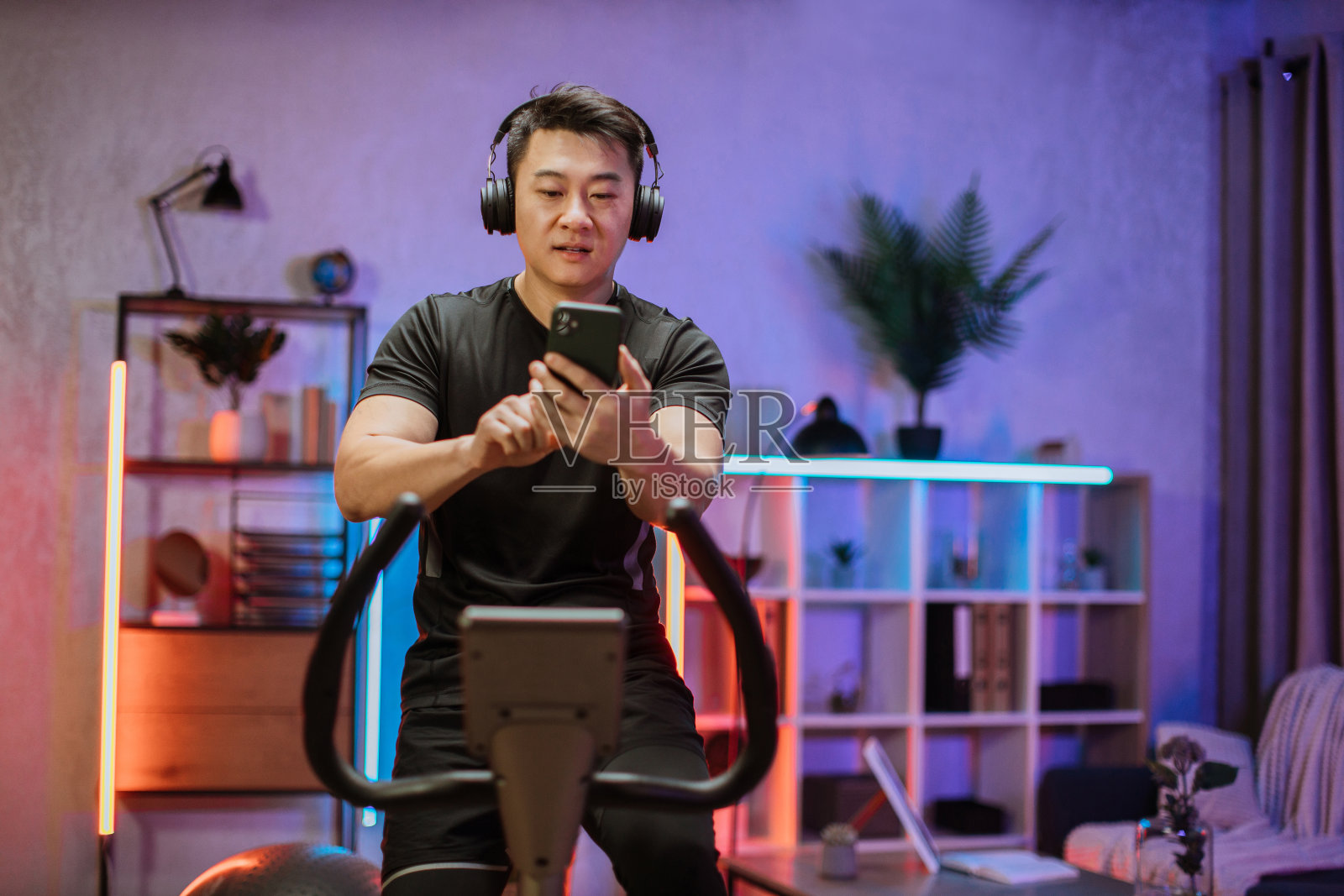 年轻的亚洲男子带着耳机在家里或健身中心的固定自行车上锻炼。照片摄影图片
