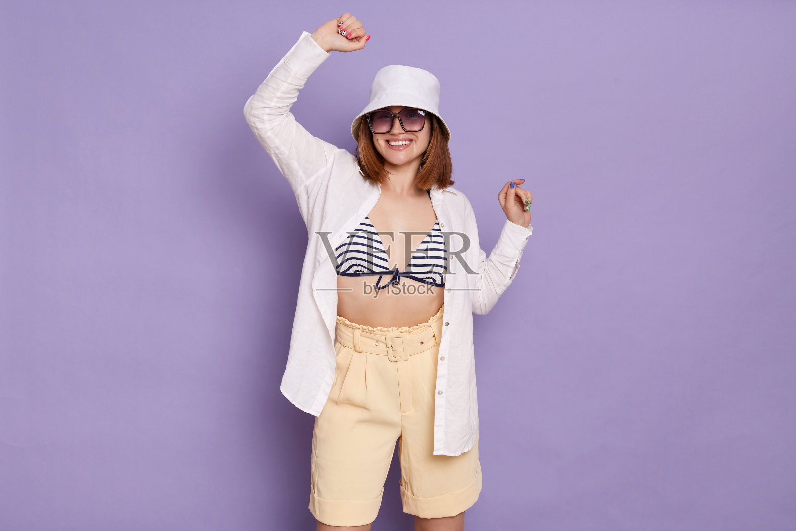 画像兴奋的女人戴着太阳镜，衬衫，巴拿马，条纹泳衣和短裤，举起手臂，庆祝她的成功，摆姿势孤立在紫丁香背景。照片摄影图片