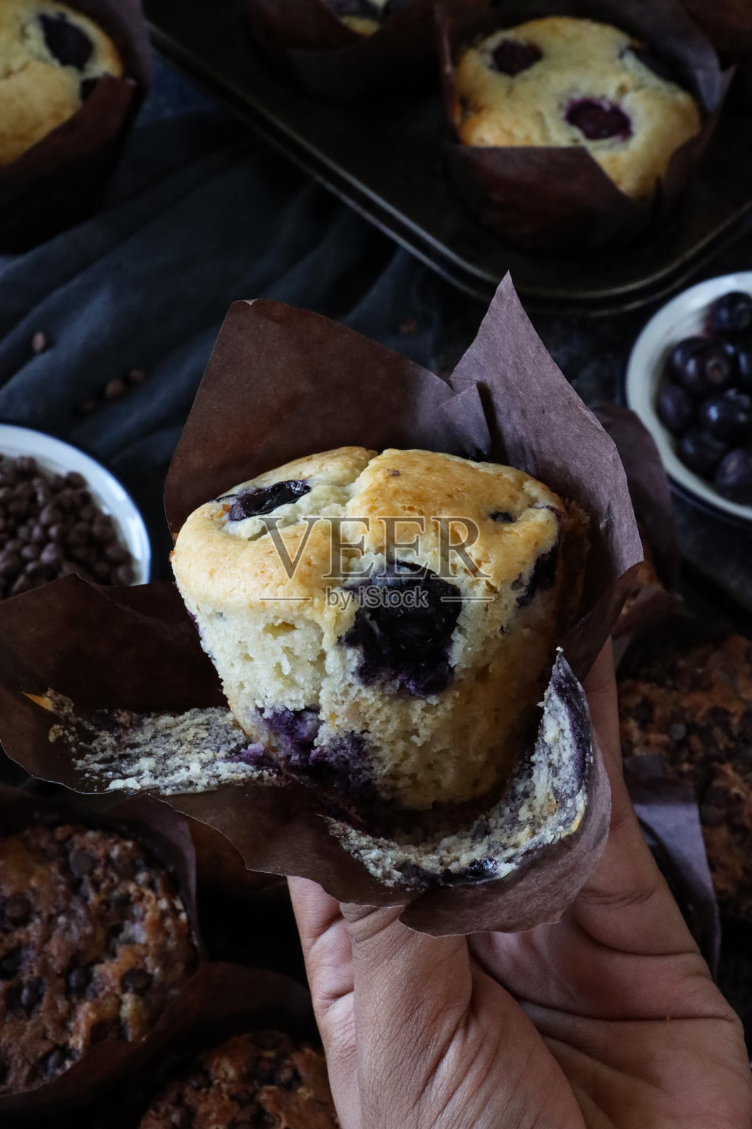 无法辨认的人抱着个人，新鲜烘焙的自制蓝莓松饼的形象，从棕色纸蛋糕盒，一碗巧克力片，蛋糕锡与烘烤上的污垢，灰色的薄纱，重点在前景照片摄影图片