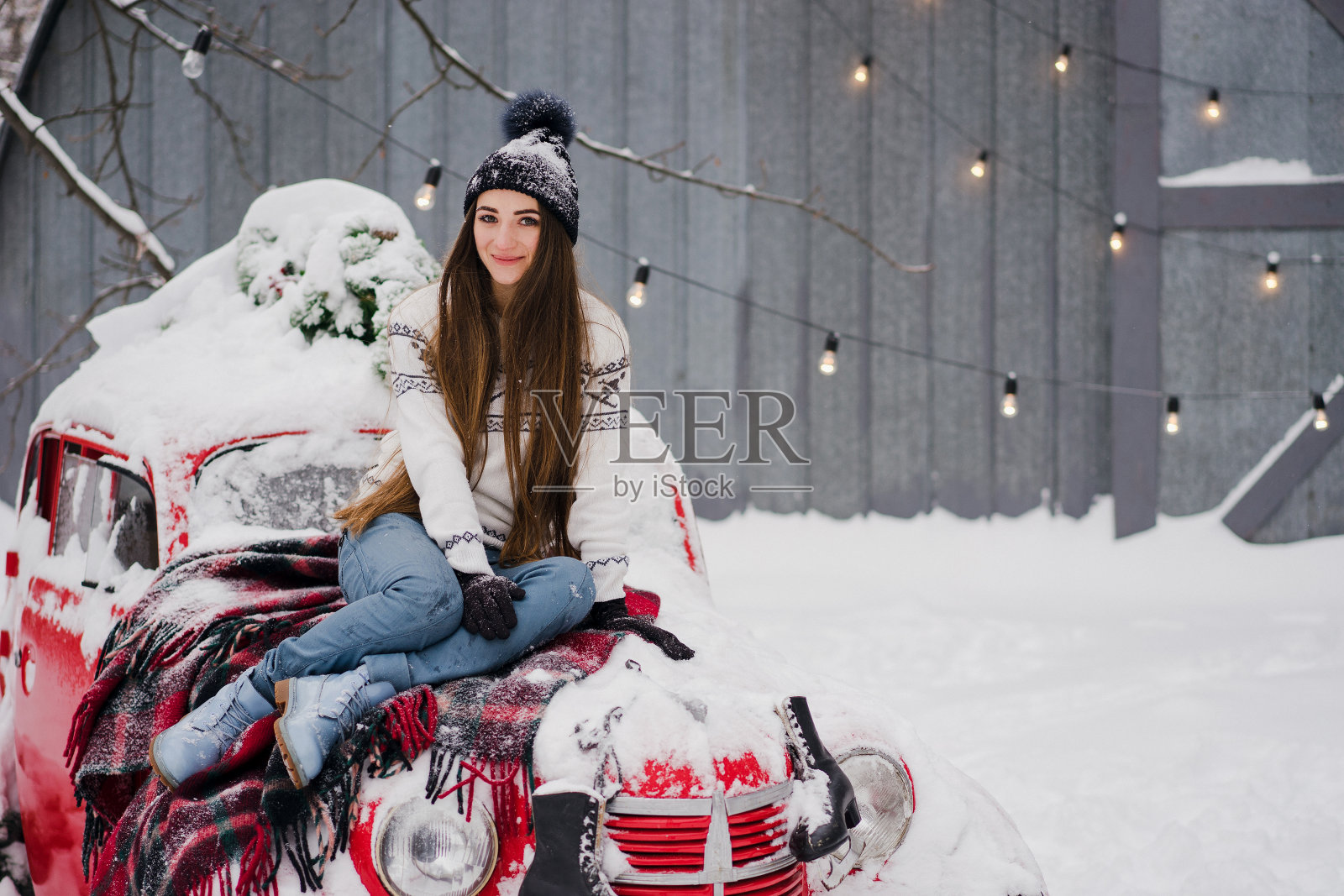 美丽的女人穿着温暖舒适的衣服坐在一辆覆盖着雪的红色老爷车上。照片摄影图片