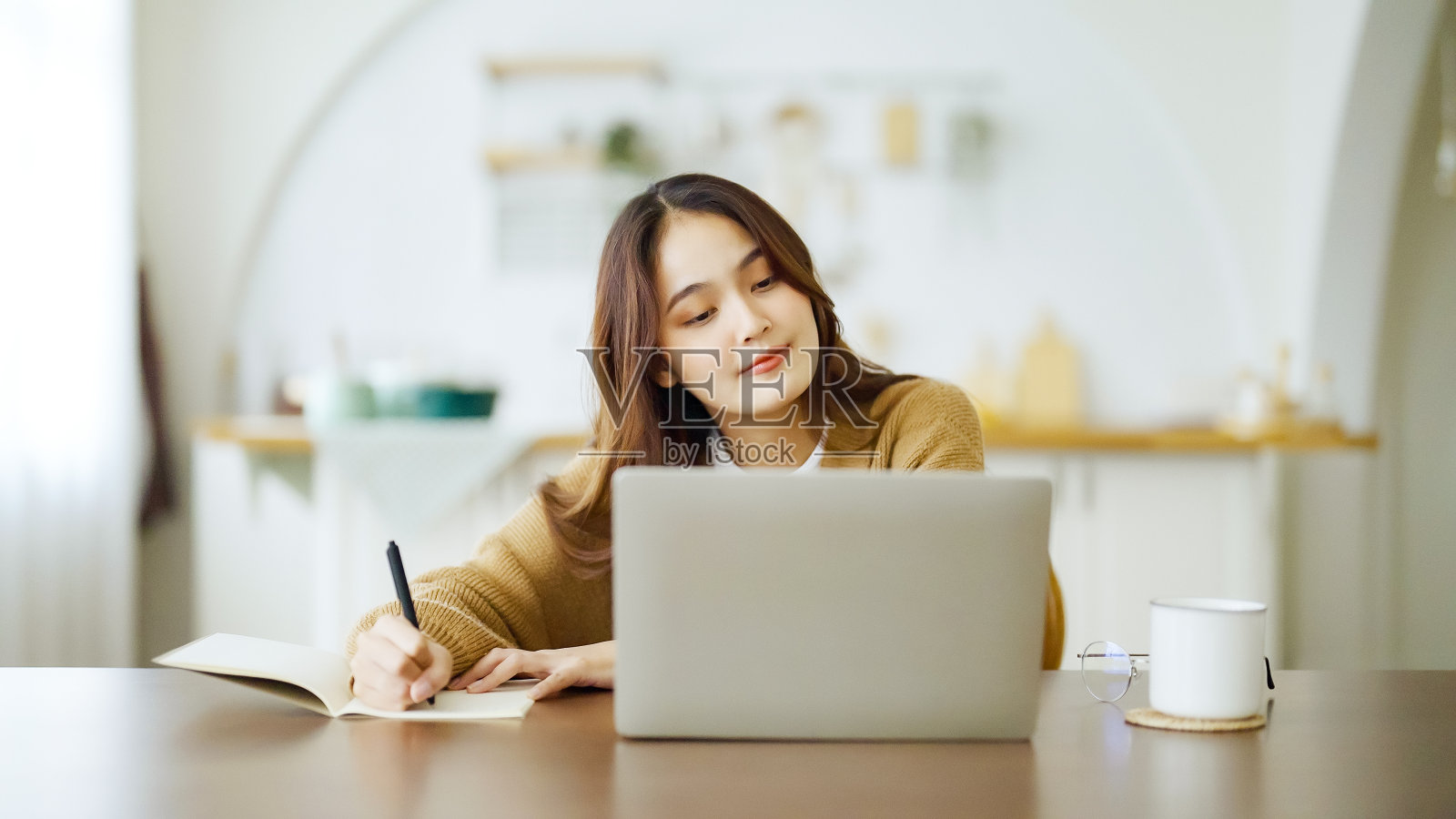 年轻的亚洲妇女坐在桌子上，在笔记本上做笔记。在桌子上用笔记本电脑工作，文书工作和一杯咖啡。学生学习在网上照片摄影图片