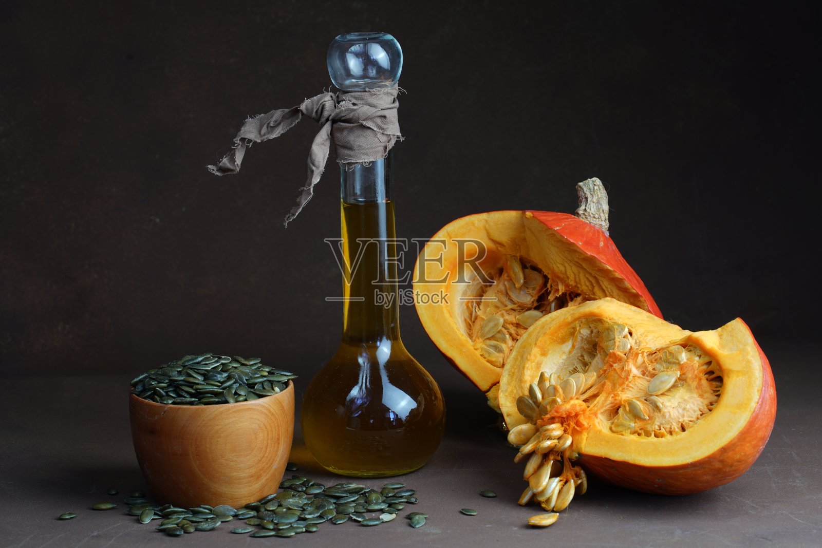静物画是用装着两半南瓜的装饰瓶里的葵花籽油和放在桌子上黑色背景上的晒过壳的油炸黑南瓜籽做成的照片摄影图片