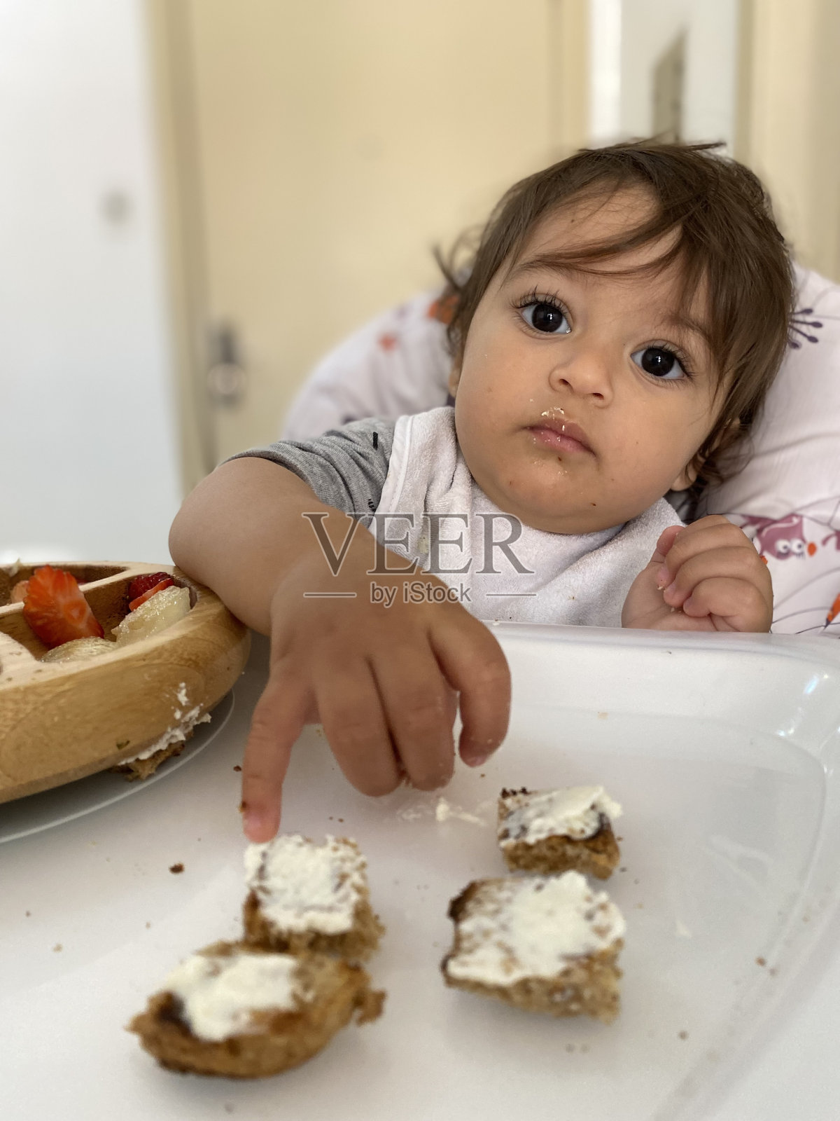 婴儿坐在喂食椅上吃他的早餐竹盘草莓，香蕉，桃子和乳清干酪全麦面包吐司。提及孩子的健康饮食，父母对孩子的照顾。照片摄影图片