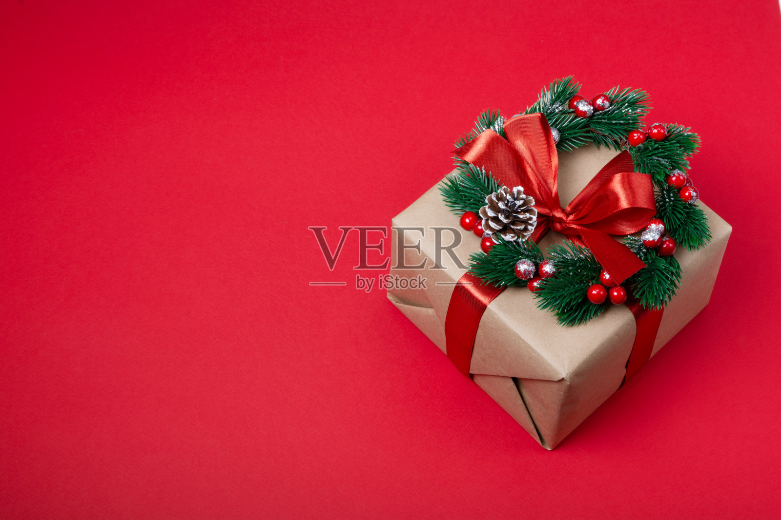 一份用牛皮纸包装的礼物，用红丝带系着，红色背景上有一个圣诞花环。新年和圣诞节的概念照片摄影图片