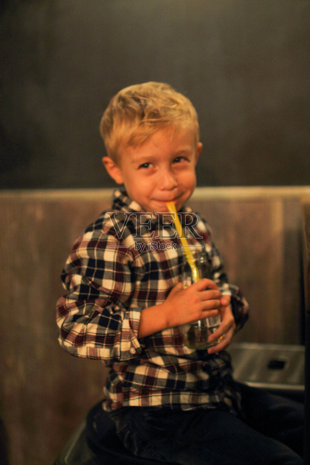 穿着格子衬衫的可爱的金发小男孩正在客厅里用吸管喝果汁照片摄影图片