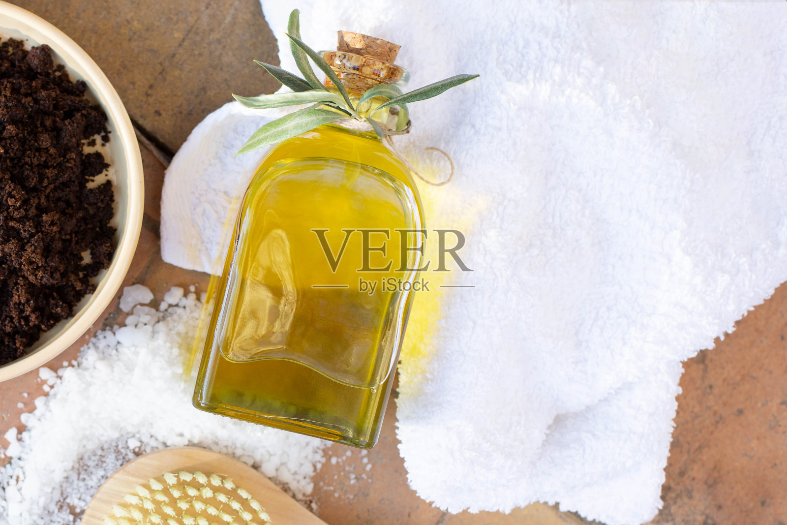 白色的浴巾上放着一瓶橄榄油，一罐手工制作的咖啡磨砂膏和身体刷。照片摄影图片