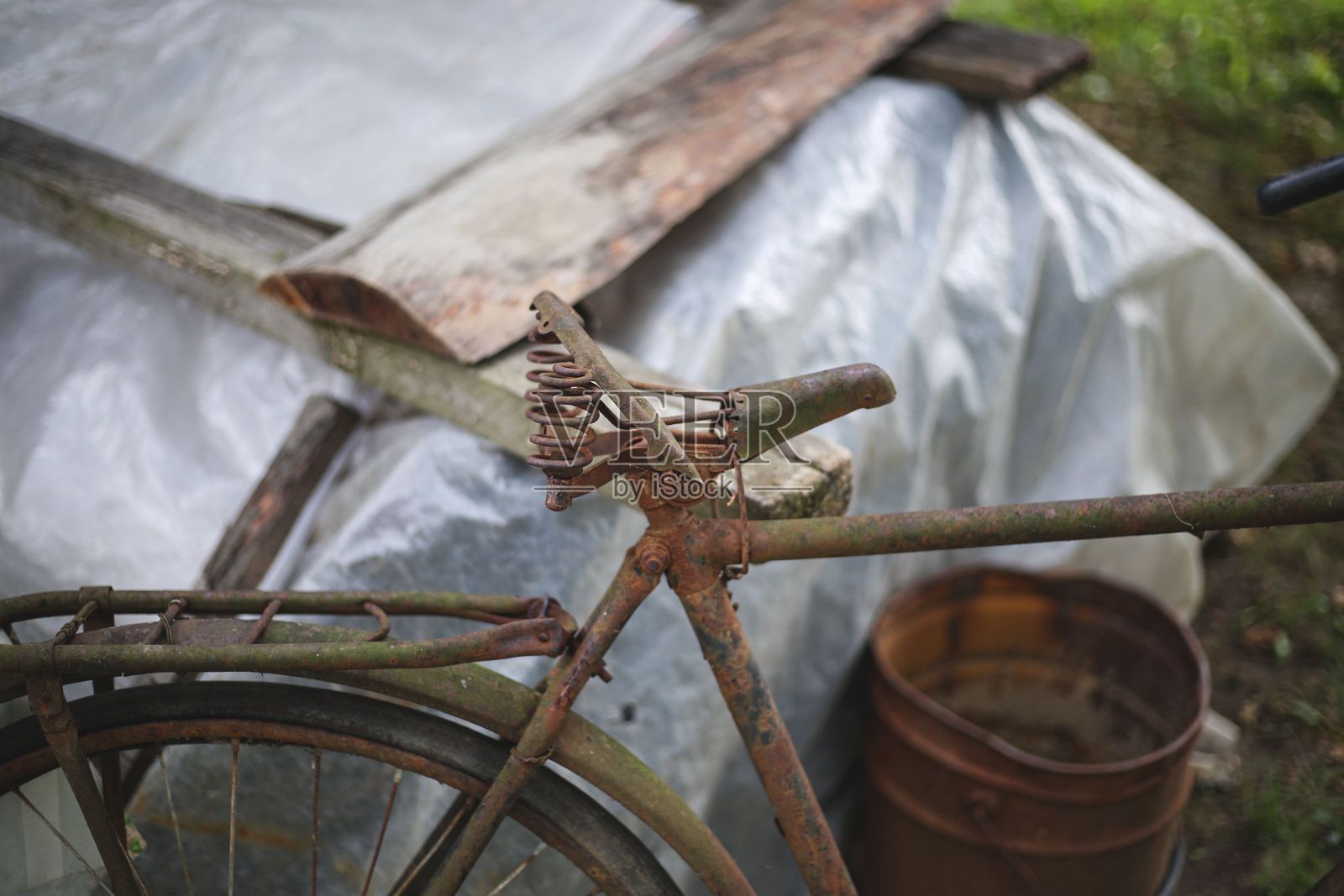 一辆锈迹斑斑的棕色金属自行车停在花园里的一个水桶旁边照片摄影图片