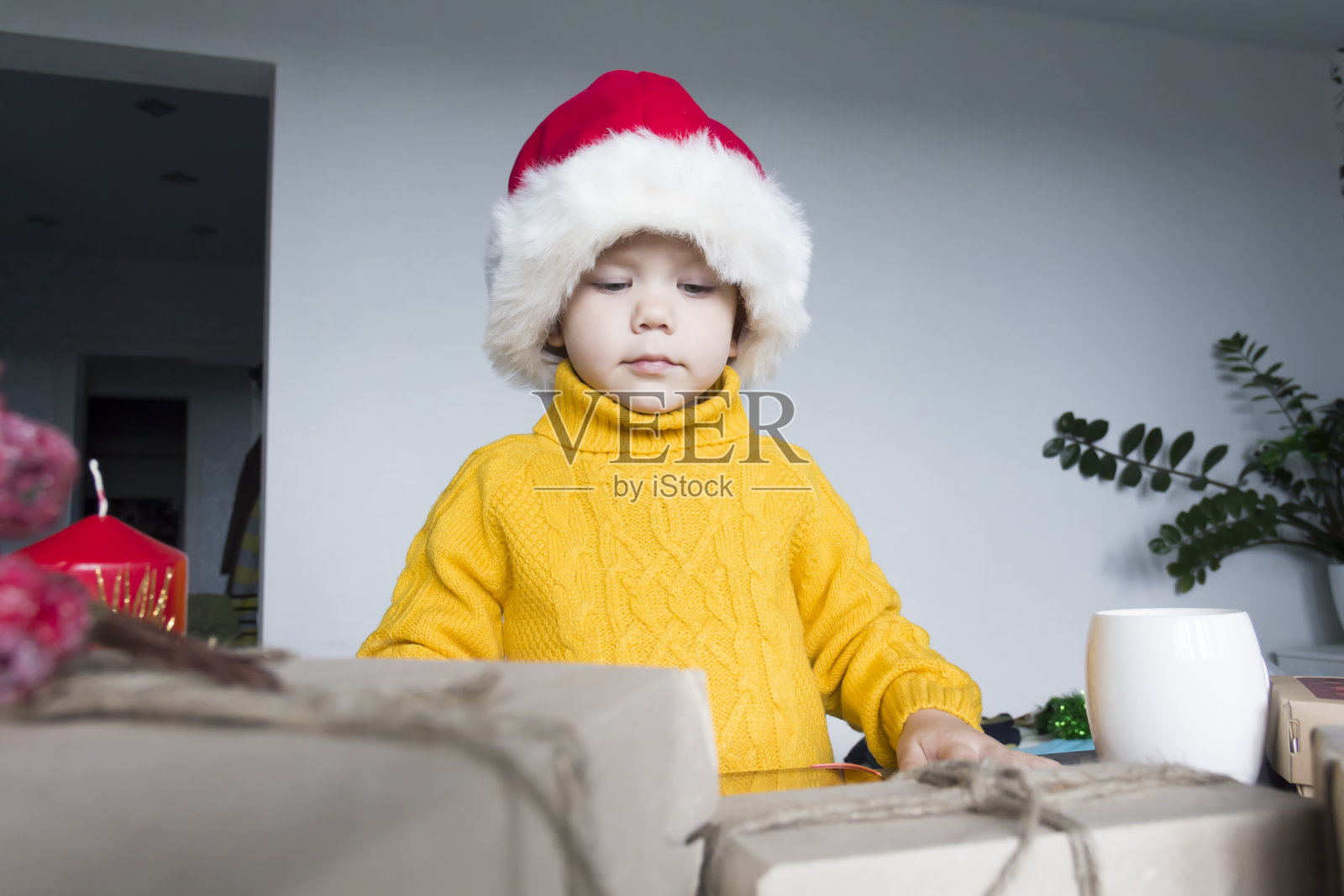 一个穿着黄色毛衣、头戴红色圣诞老人帽的可爱小男孩坐在一张木桌旁，桌上放着用牛皮纸包装的礼物。照片摄影图片