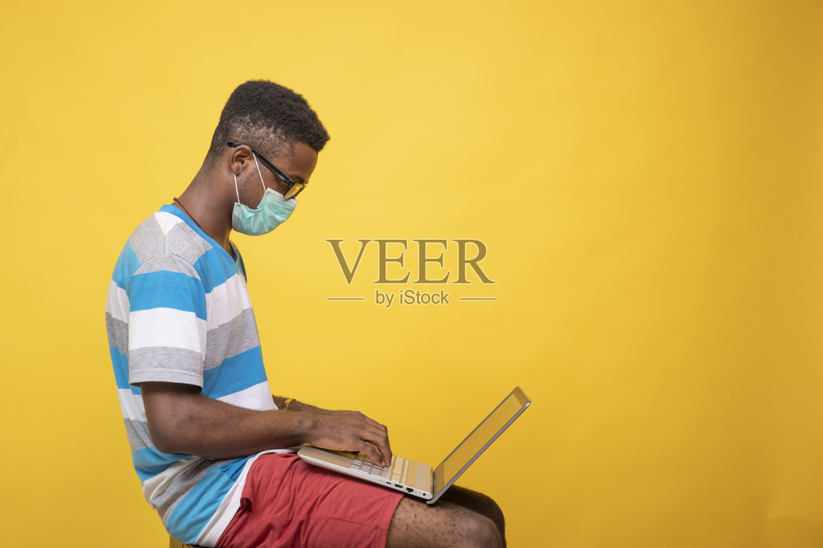 年轻的非洲男性戴着眼镜和口罩在笔记本电脑上工作- COVID-19照片摄影图片