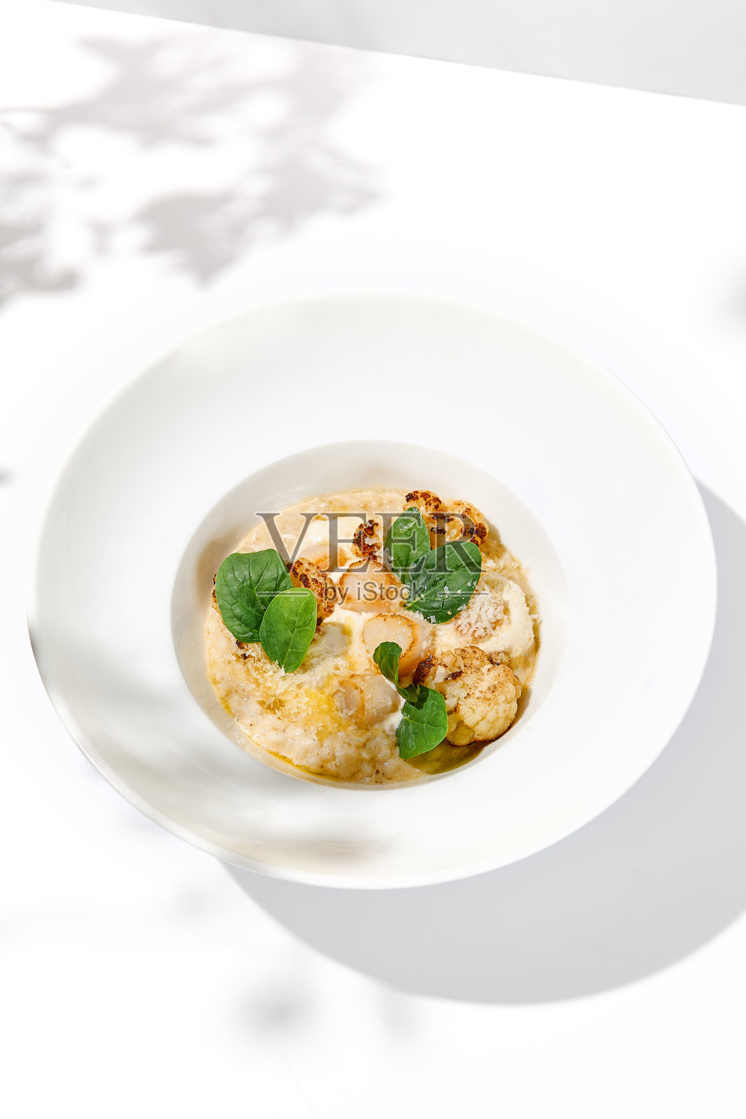 海鲜烩饭配扇贝和菜花，装在白色盘子里。奶油烩饭配海贝和花椰菜。意大利烩饭与扇贝在光背景与树叶阴影照片摄影图片