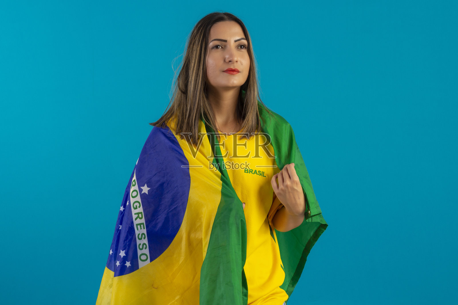巴西女支持者，穿着印有巴西国旗的衬衫。巴西球迷在工作室的照片与蓝色背景和巴西的颜色。照片摄影图片