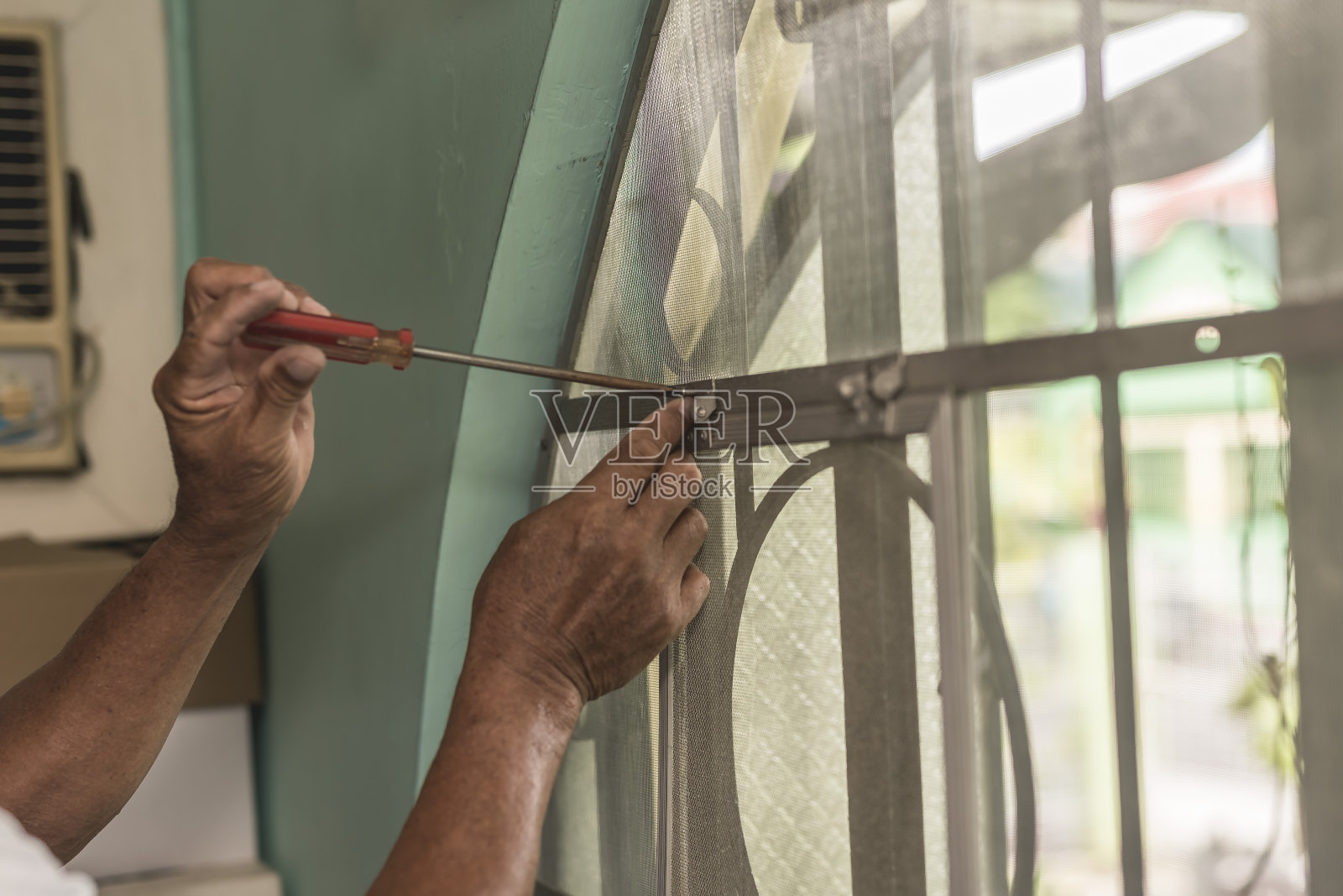 一名杂工正在卧室的窗户上用螺丝刀安装纱网框。家居装修概念。照片摄影图片