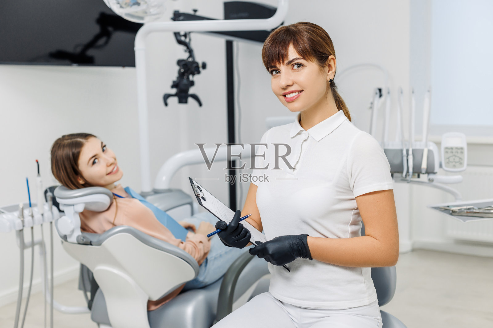 在检查牙齿时，友好的牙医将药片放在病人的背景上。现代牙科诊所。专业口腔护理理念，口腔诊室会诊。照片摄影图片