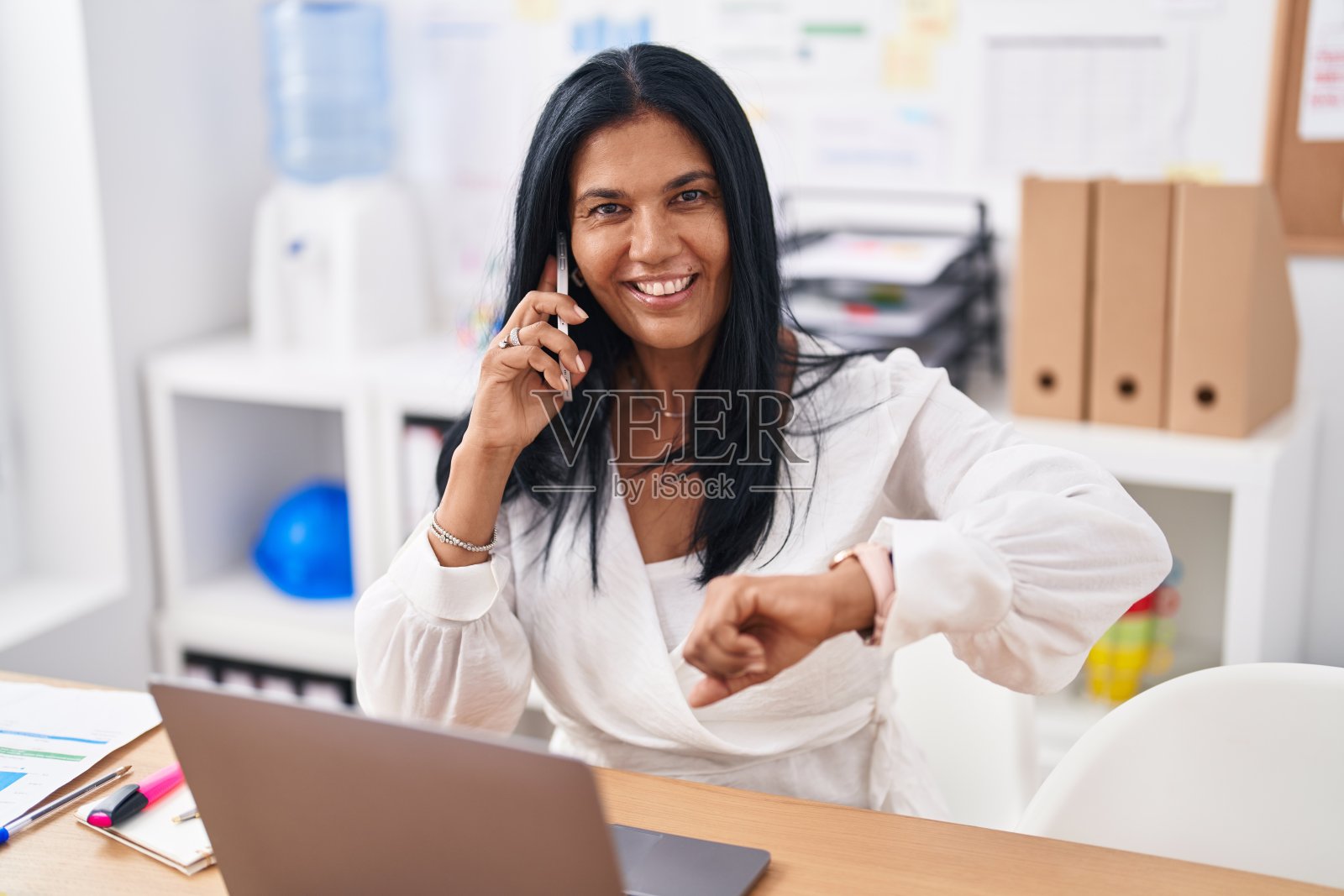 中年西班牙女性商务工作者在办公室用智能手机看手表说话照片摄影图片