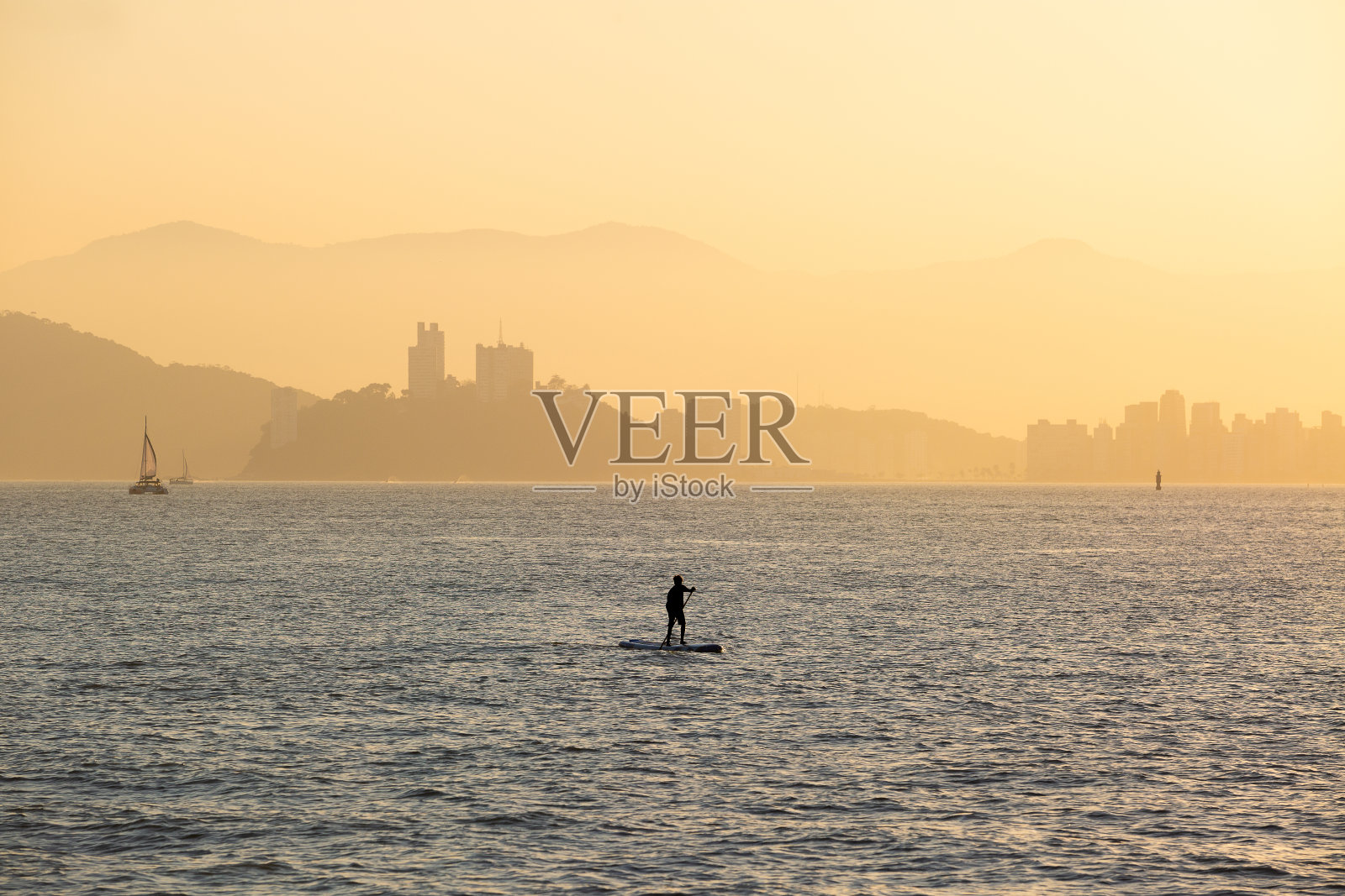 男孩在巴西圣保罗桑托斯湾练习站立划水。背景是圣维森特的波查特岛照片摄影图片