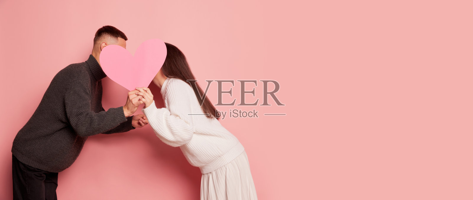 可爱的年轻夫妇的肖像，男人和女人接吻背后的纸心，摆出孤立的粉色背景。带有复制空间的传单照片摄影图片