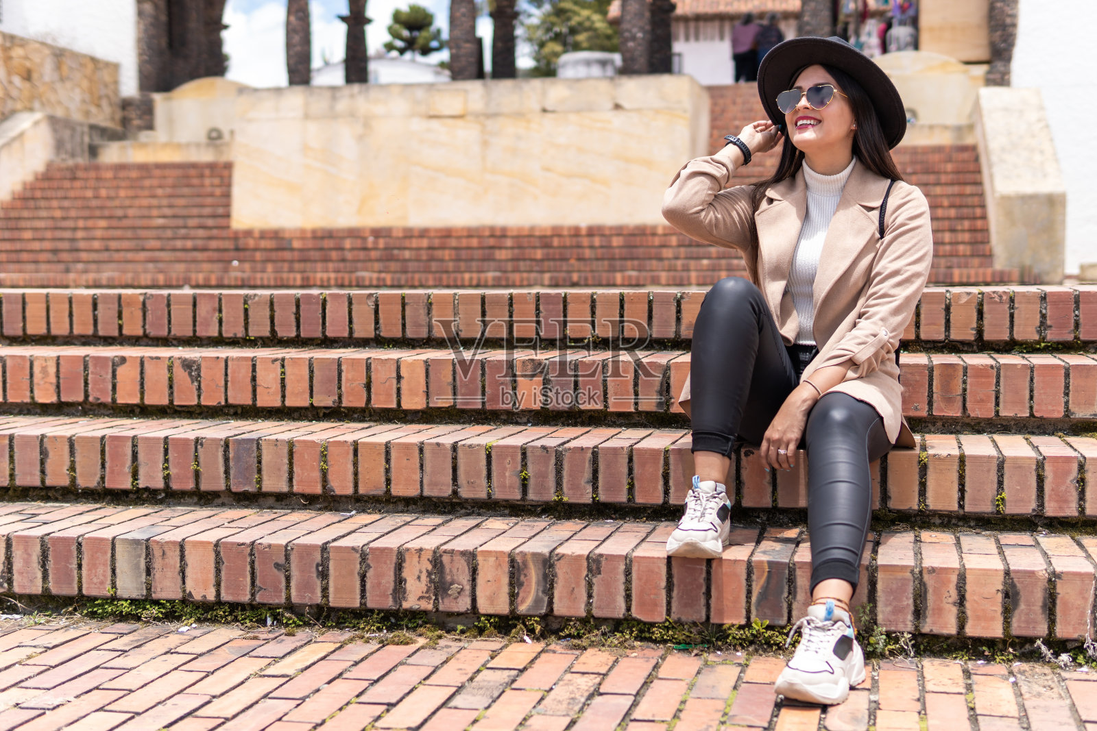 戴眼镜、穿外套、戴帽子的女人坐在广场的台阶上。照片摄影图片